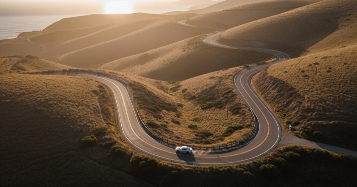 Aerial view of a ribbon of empty mountain road carving through golden hills at dawn, with a single sports car mid-corner